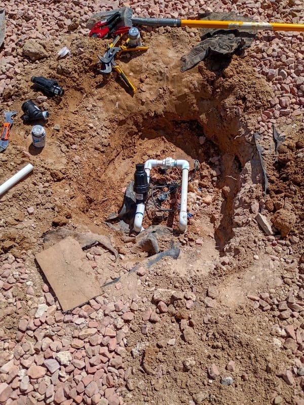 An irrigation valve exposed in an excavated trench, with PVC pipe and cutting tools laid out beside it.